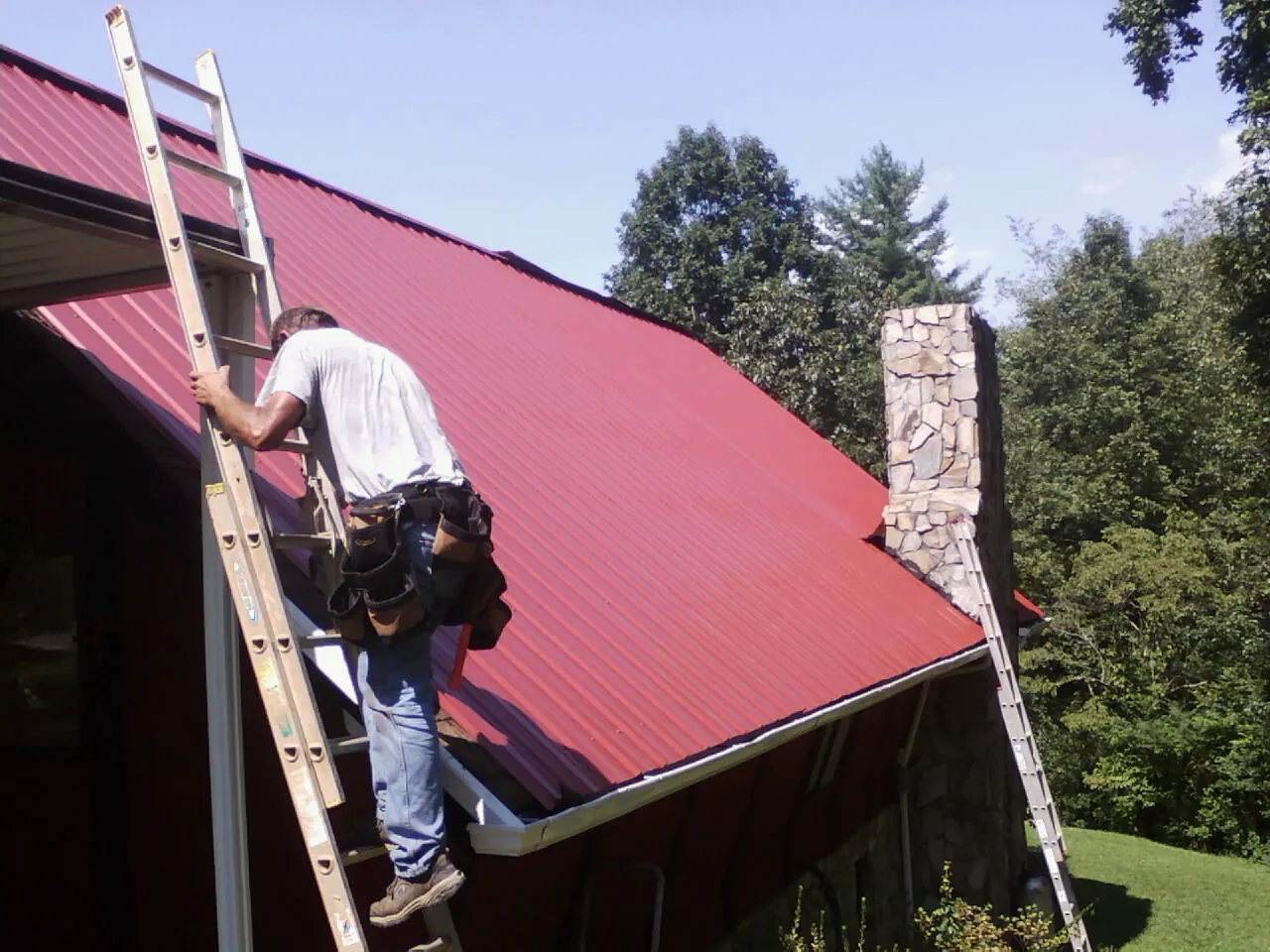 Roofer on a ladder beside a red metal roof and stone chimney — Soffit & Fascia Repair by Roofing Brothers in Honey Creek